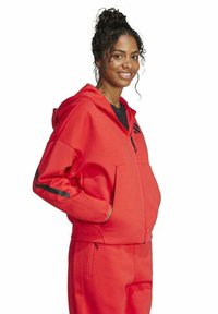 Eine Frau lächelt, trägt einen roten Reißverschluss-Hoodie und passende rote Hosen, die Hände in den Hoodie-Taschen, lockiges Haar zusammengebunden.