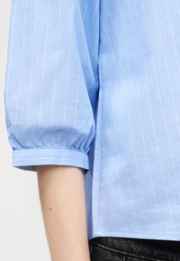 Ανοιχτό μπλε πουκάμισο με ριγέ σχέδιο και κοντά φουσκωτά μανίκια που φτάνουν λίγο πάνω από τον αγκώνα, φορεμένο με μαύρο παντελόνι.