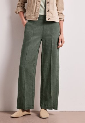 Femme portant un pantalon large taille haute vert olive, des chaussures beiges en suède faciles à enfiler, un top vert clair et une veste beige boutonnée aux manches retroussées.