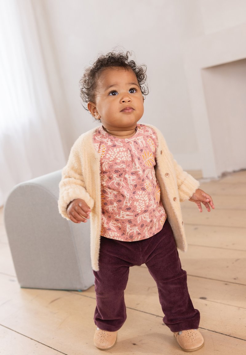 Ein Kind trägt eine hellbeige, flauschige Strickjacke über einem rosa gemusterten Hemd und dunkellila Cordhosen, kombiniert mit beigen Schuhen.