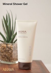 Vit tub av AHAVA Mineral Shower Gel med brunt lock. Texten lyder "VATTEN" och "200 ml." Mot en suddig bakgrund av grönska.