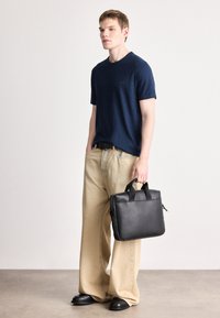 Μαύρη δερμάτινη τσάντα με διπλές λαβές, λεία υφή, ορθογώνιο σχήμα. Το μοντέλο φοράει μπλε σκούρο μπλουζάκι και μπεζ φαρδιά παντελόνια.