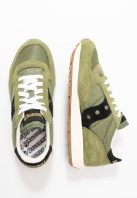 Vihreät mokkanahasta ja verkkomateriaalista valmistetut urheilukengät, joissa on valkoiset nauhat, mustat yksityiskohdat ja kontrastivärinen kumipohja. Logo on kengän kielessä.