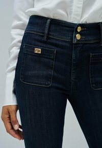 Jeans de color azul oscuro con una cintura alta, que cuentan con dos botones dorados, bolsillos frontales y un acento decorativo en el bolsillo trasero.