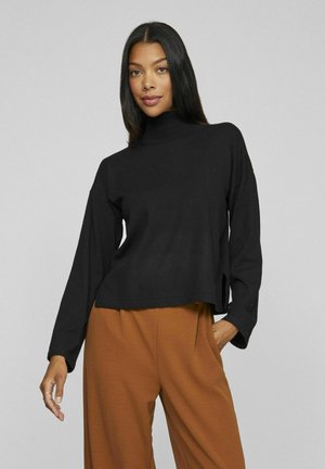 Maglia nera a maniche lunghe con collo a lupetto, dal taglio morbido e con spacchi laterali, abbinata a pantaloni beige a gamba larga. Design minimalista e texture morbida.