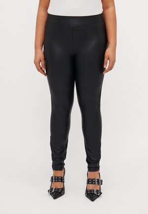 Person iført sorte leggings i faux læder og sorte spidse hæle med dobbelte spænder, stående mod en hvid baggrund.