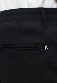 Gros plan sur la ceinture d'un pantalon noir avec passants de ceinture, poche arrière et petit logo argenté en forme de "R".