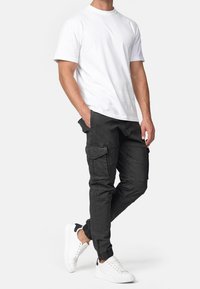 Man in een eenvoudig wit T-shirt met korte mouwen, zwarte cargobroeken met zakken en ritsen, en witte sneakers met een zwart hieldetail.