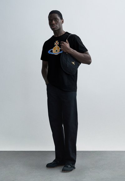 Μαύρο t-shirt με γραφικό σχέδιο σε κίτρινο και μπλε, συνδυασμένο με σκούρο τζιν παντελόνι και μαύρα παπούτσια. Το μοντέλο κρατάει μια μαύρη τσάντα.