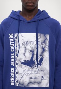 Μπλε φούτερ από βαμβάκι με γραφικό σχέδιο μιας αετού και κείμενο. Περιλαμβάνει κουκούλα με κορδόνι και επιχρυσές αλυσίδες.