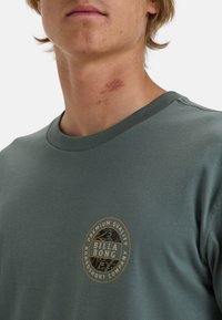 Grünes Baumwoll-T-Shirt mit kurzen Ärmeln, rundem Ausschnitt und einem runden Logo auf der linken Brust, das Text und ein detailliertes Muster zeigt.