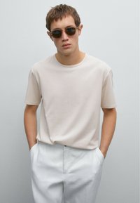 Light beige short-sleeve t-shirt made from soft material, paired with white trousers. Model wears round sunglasses. Neutral background.