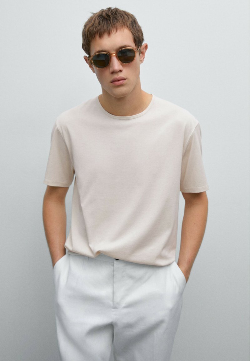 Light beige short-sleeve t-shirt made from soft material, paired with white trousers. Model wears round sunglasses. Neutral background.