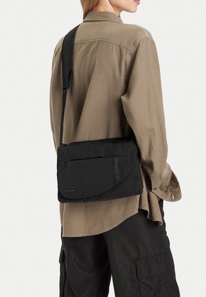 Osoba v světle hnědé košili a černých cargo kalhotách, nesoucí černou crossbody tašku značky G-Star přes rameno, otočená zády.