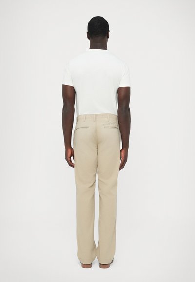 Calças bege de corte reto com um brilho subtil e dois bolsos nas costas. Combinadas com uma t-shirt branca justa, vistas de trás.