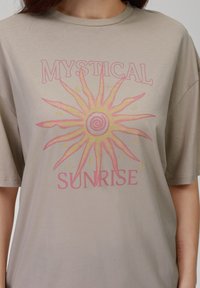 Lys beige T-skjorte med et grafisk soldesign i rosa og gult med teksten "MYSTISK SOLOPPGANG." Korte ermer og rund hals.