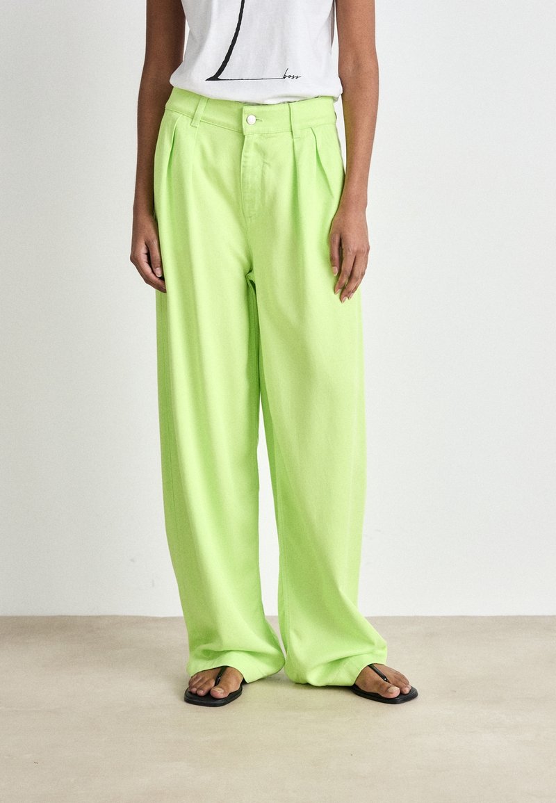 Personne portant un pantalon ample taille haute vert citron associé à un t-shirt blanc à motif et des tongs noires, debout sur un sol clair.
