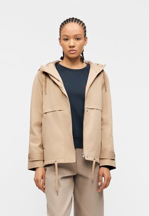 Joven con cabello trenzado que lleva una chaqueta con capucha beige sobre una camiseta azul oscuro y pantalones beige, de pie frente a un fondo liso.