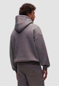 Felpa grigia con un ampio cappuccio, maniche larghe e una texture morbida. Indossata con pantaloni grigi abbinati, caratterizzati da un dettaglio sulla tasca posteriore.