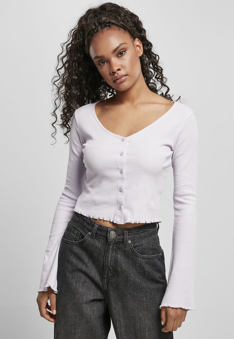 Svetle levandulový cropped top s V-výstřihem, knoflíkovým zapínáním a třepeným lemem, doplněný o tmavě šedé džíny s vysokým pasem. Dlouhé rukávy se zvonovými konci.
