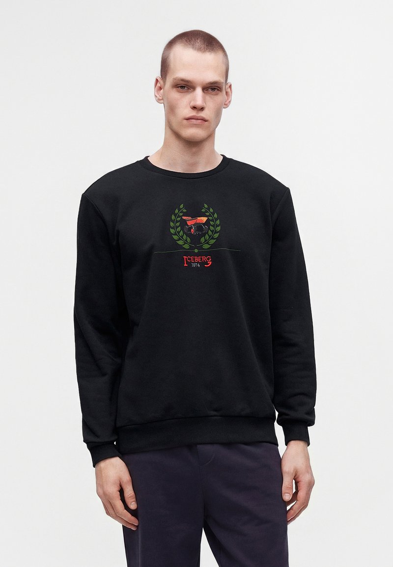 Jeune homme aux cheveux courts portant un sweat-shirt noir avec une couronne de laurier verte et un logo d'avion rouge, debout devant un fond blanc uni.