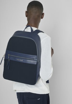 Homme portant un pull blanc et un sac à dos bleu marine avec une poche zippée à l'avant, debout dos à la caméra devant un fond gris uni.