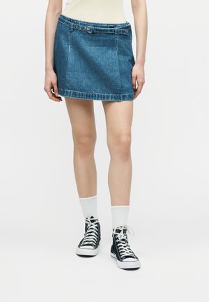 Άτομο που φοράει μπλε μίνι φούστα από τζιν με θηλιές για ζώνη, λευκές κάλτσες μέχρι τον αστράγαλο και μαύρα ψηλά αθλητικά παπούτσια, στέκεται μπροστά σε μια μονόχρωμη επιφάνεια.