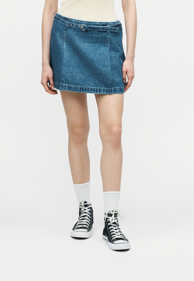 Άτομο που φοράει μπλε μίνι φούστα από τζιν με θηλιές για ζώνη, λευκές κάλτσες μέχρι τον αστράγαλο και μαύρα ψηλά αθλητικά παπούτσια, στέκεται μπροστά σε μια μονόχρωμη επιφάνεια.