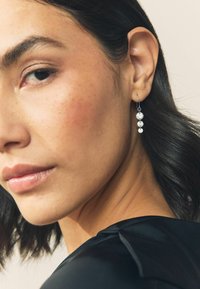 Lähivõte naisest, kes kannab musta ülaosa ja hõbedasi tilk-kujulisi kõrvarõngaid, millel on kolm ümarat, läbipaistvat vääriskivi.