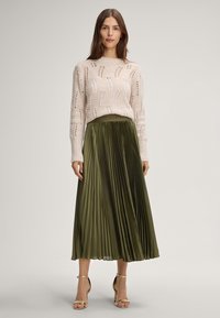Woman wearing a cream knit sweater with patterns, olive green pleated midi skirt, and gold high-heeled sandals, standing on a light background.