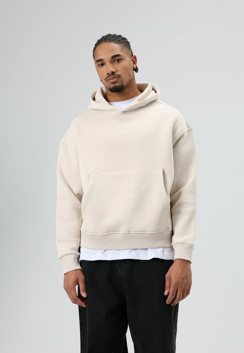 Beige sweatshirt met capuchon, grote zak aan de voorkant, losse pasvorm en geribbelde boorden. Draagt over een wit T-shirt, gecombineerd met donkere spijkerbroek.