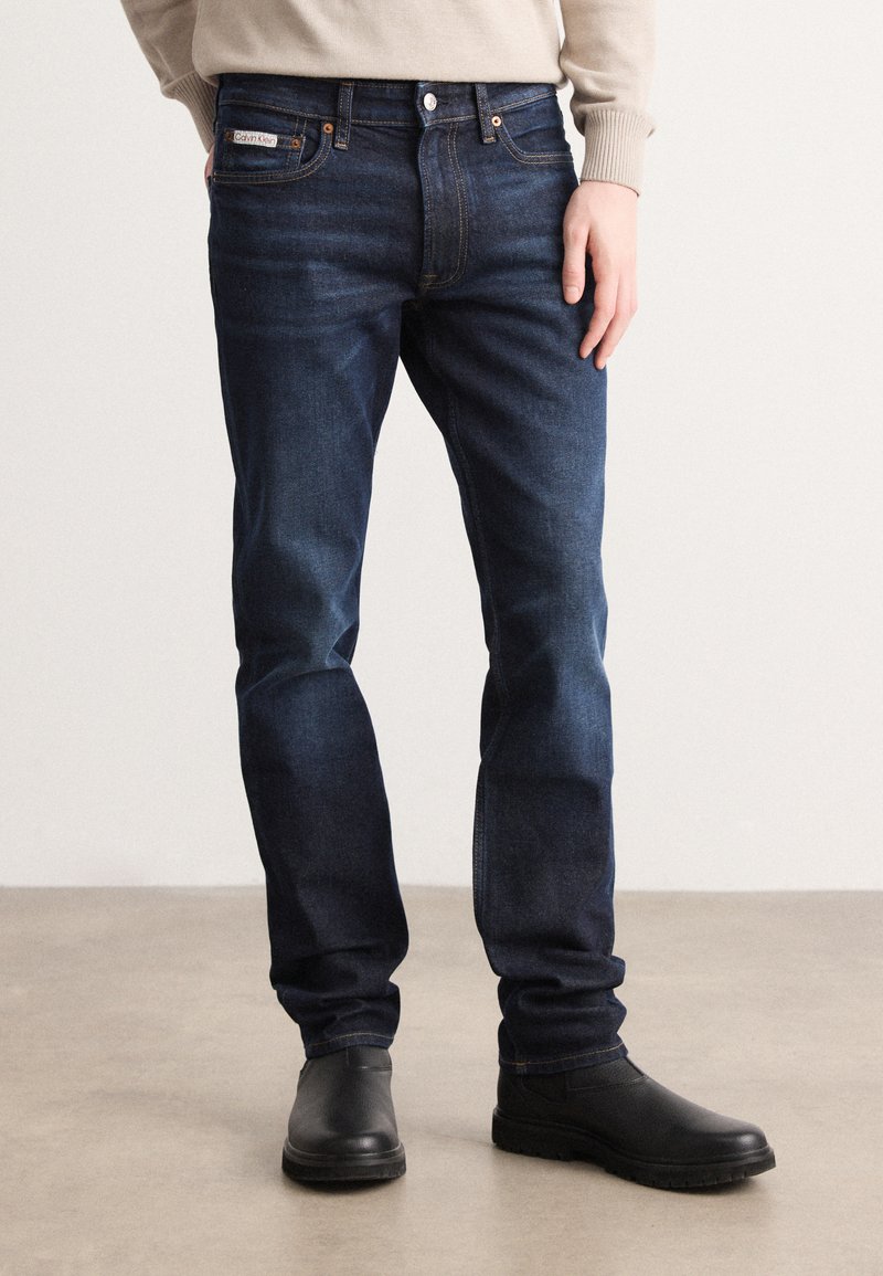 Mørkeblå denimjeans med rett passform, fem-lomme-design og detaljer i bronsefarge. Kombinert med svarte ankelstøvler.