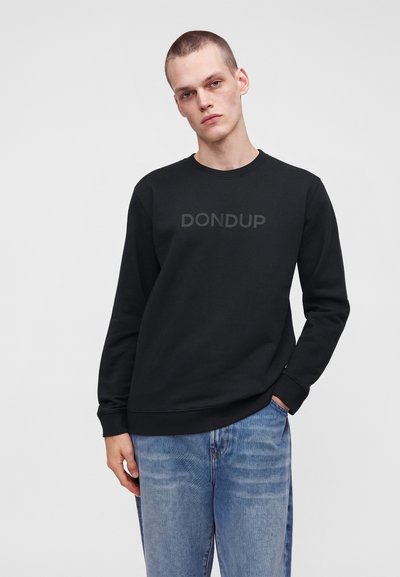 Jeune homme aux cheveux courts portant un sweat-shirt noir avec le texte "DONDUP" et un jean bleu clair sur un fond uni clair.