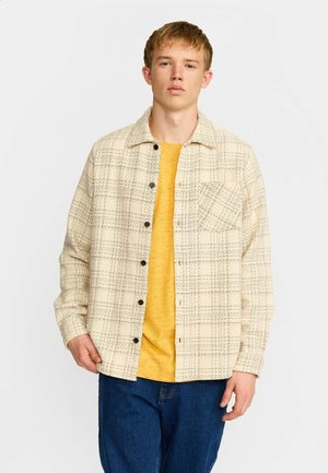 Jeune homme portant une chemise à carreaux crème boutonnée sur un t-shirt jaune et un jean bleu, debout devant un fond uni clair.
