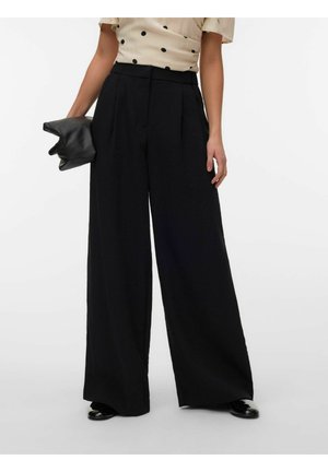 Kvinne iført svarte, vidbeinte bukser med høyt liv, kremfarget bluse med svarte polkadotter, holder en svart clutch i venstre hånd.