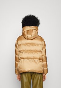 Giacca puffer imbottita marrone con cappuccio, caratterizzata da una texture lucida, sezioni cucite e un taglio rilassato, abbinata a pantaloni verdi.