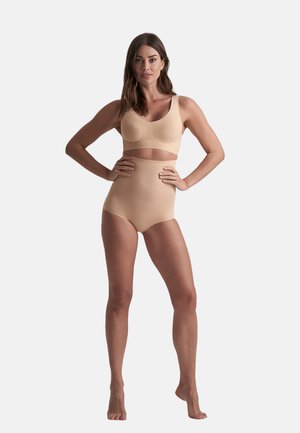 Vrouw die blootsvoets staat met handen op de heupen, gekleed in naadloze huidkleurige high-waist shapewear slip en bijpassende bh tegen een effen achtergrond.