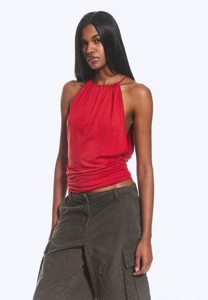Donna con lunghi capelli neri che indossa un top rosso senza maniche con collo all'americana e pantaloni cargo verde scuro, in piedi su uno sfondo semplice.