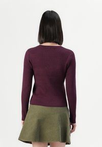 Femme aux cheveux noirs et lisses portant un pull violet à manches longues côtelé ajusté et une jupe évasée vert olive, debout, de dos.