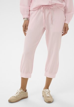 Žena stojaca na bielom pozadí, oblečená v svetloružových krátkych športových nohaviciach so sťahovacou šnúrkou a béžových teniskách.