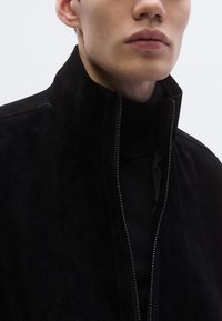 Μαύρο σακέτο από σουέντ με ψηλό γιακά και φερμουάρ, με ανάγλυφη υφή και εφαρμοστή γραμμή, φορεμένο πάνω από μαύρο πουλόβερ με ζιβάγκο.