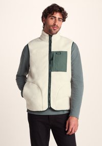 Fleece vest in cream with a green chest pocket. Features a high collar, front zip, and large side pockets. Textured fabric and layered design.