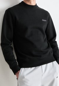 Schwarzer Sweatshirt mit Rundhalsausschnitt, gerippten Ärmelbündchen und Saum, mit einem kleinen gestickten Logo auf der Brust. Aus glattem Baumwoll-Mischgewebe.
