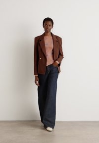 Brun skreddersydd blazer med gullknapper, lyseblå topp og vide mørk denimjeans. Modellen har på seg hvite sko og står på en lys bakgrunn.