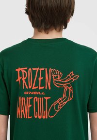 Roheline T-särk, millel on oranž graafiline tekst "FROZEN WAVE CULT" koos käe ja jala kujutisega. Lühikesed varrukad ja ümar kaelus. Sujuv kangastekstuuri.
