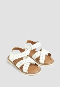 Sandali bianchi in pelle con bordi scallopati e motivi floreali perforati. Suola piatta, design aperto e cinturini regolabili per una vestibilità sicura.