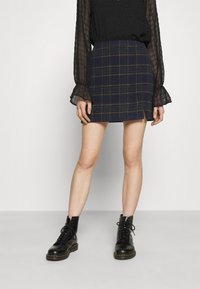 Marinblå rutig minikjol med gröna och gula accenter, tillverkad av ett texturerat tyg. Matchas med svarta snörstövlar. Blus med lång ärm i genomskinligt material.