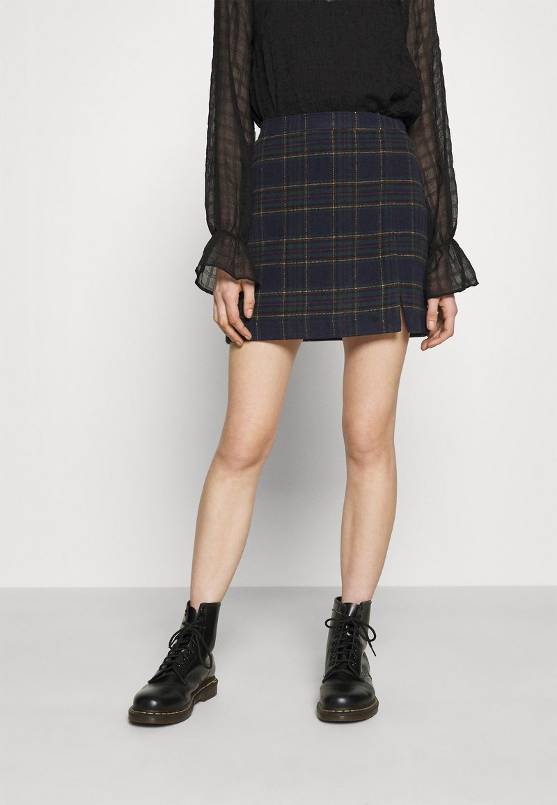 Marinblå rutig minikjol med gröna och gula accenter, tillverkad av ett texturerat tyg. Matchas med svarta snörstövlar. Blus med lång ärm i genomskinligt material.