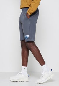 Pantaloni sportivi grigi realizzati in tessuto traspirante, con vita elastica, tasche laterali e una piccola toppa grafica sulla coscia.