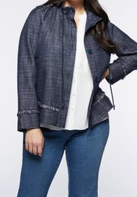 Marinblå texturerad jacka med fransad kant, knappar fram och dragsko. Bärs över en vit topp, kombinerad med blå jeans.